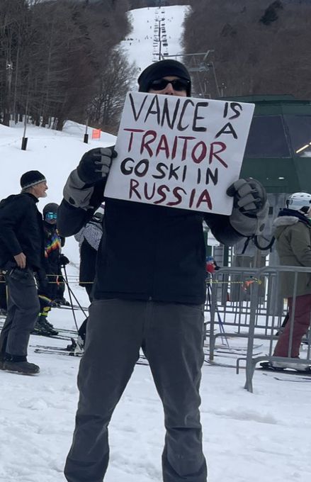 A person stands in a ski area holding a sign that reads, "VANCE IS A TRAITOR GO SKI IN RUSSIA," amid other skiers and trees.