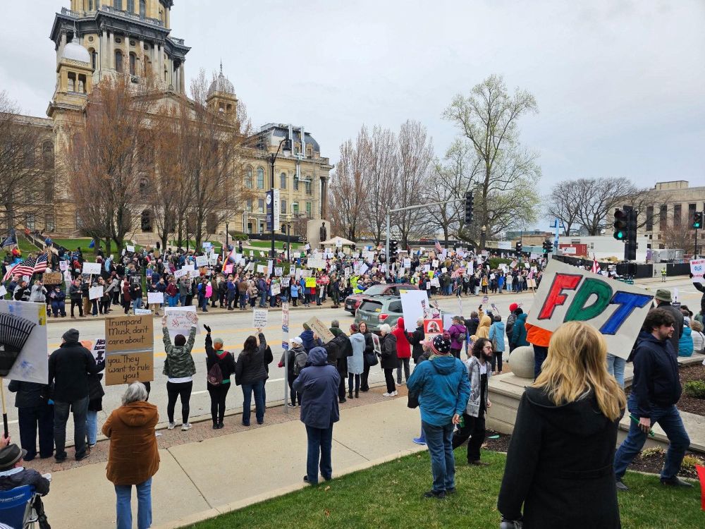 A large group of protesters gather outside a historic building, holding signs and flags, advocating for justice and liberty on a cloudy day.