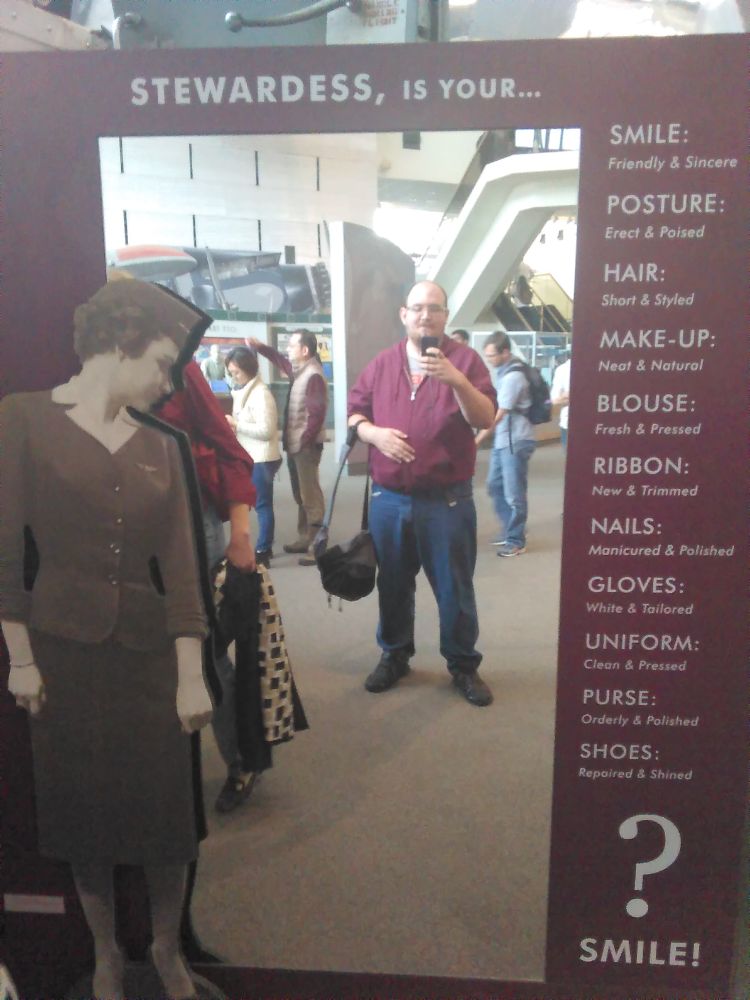 Photo of a pre-transition Susan (white egg with beard and glasses wearing a burgundy hoodie and baggy jeans, carrying a backpack as if it were a purse) at the Smithsonian Air & Space Museum, circa 2015. She is taking a selfie in front of a mirror asking sexist early-to-mid 20th century questions to ensure that I, a male-presenting Narnian transfeminine egg, would make a good stewardess. A giant black & white photo of a mid-20th century flight attendant is besides the left frame of the mirror.