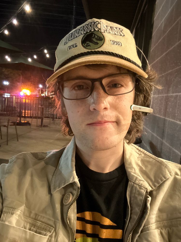 picture of a white femme dressed as a jurassic park tour guide. she is on her smoke break smiling plainly. her costume consists of a khaki jacket, houston museum of natural science shirt, a jurassic park staff hat, glasses, and a headset.