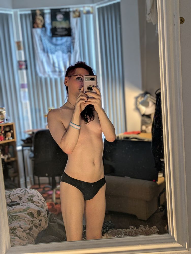 A selfie of Luci stood in front of a mirror in her underwear. She's lit from the front in warm tones, and her phone covers half her face while her one visible eye is looking directly into the camera.
The room around her looks warm, cosy, and inviting. There is the corner of a bed visible to her left, a comfortable if well used fabric armchair to her right and behind her. A shelf and desk are visible in the background though their contents are blurred and too small to make out details. The window behind her runs floor to ceiling and while the blinds are closed, the cold early morning light seeps through, at odds with the warm lighting gently suffusing the rest of the room.
She looks kinda hot, too! :3