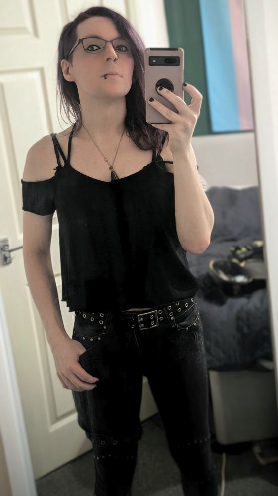 Luci taking a selfie in front of her mirror. She's wearing black jeans, a belt, and a strappy top that shows her shoulders and upper chest. She's still not smiling... She's never smiling in pictures.