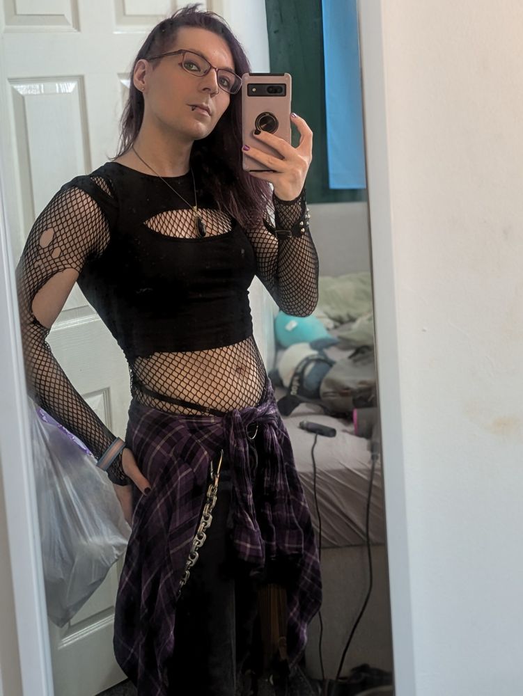 Luci stood in front of the same mirror, now with more clothes. She's wearing a tight black cropped vest with a fishnet shirt underneath, black jeans, with a purple plaid shirt tied about her waist. Her expression could perhaps best be described as "dead inside".