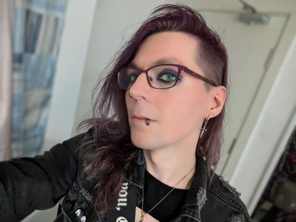 Selfie of Luci taken in the room she lives in. The background is blurred and she has purple and black eyeliner, is wearing a black jacket and still doesn't know what face to pull for pictures. 