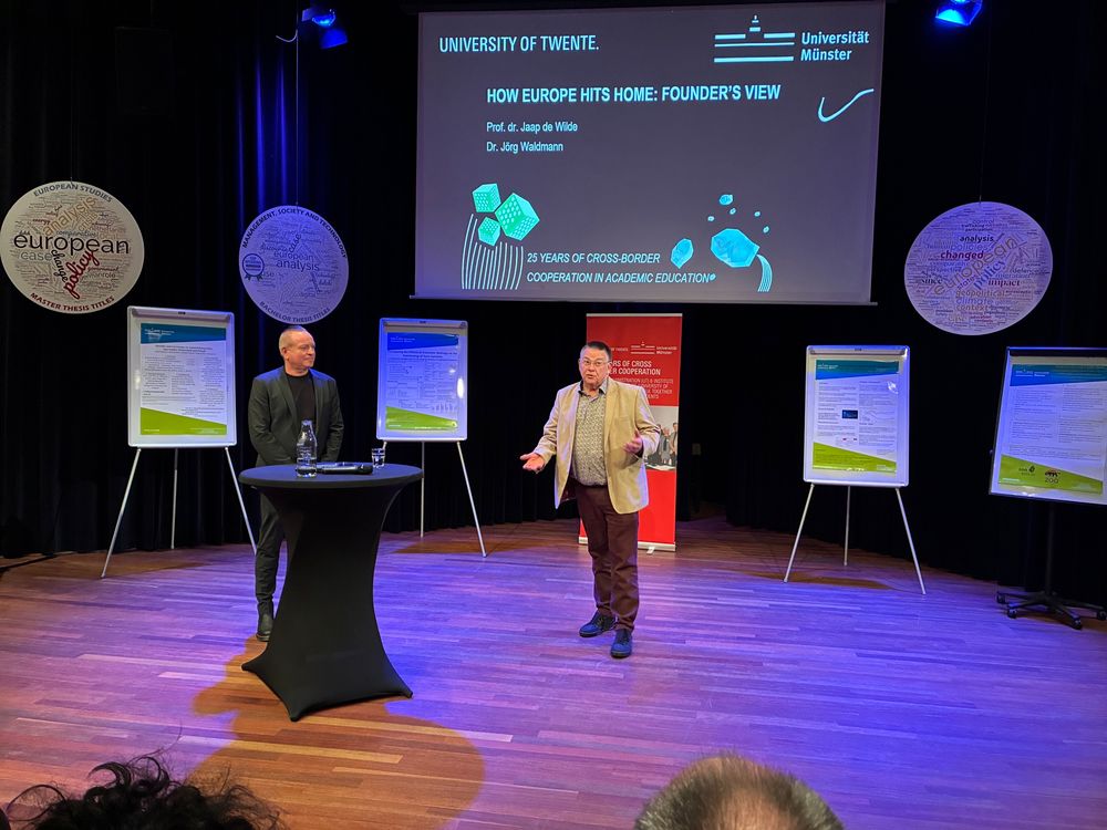Foto van Jörg Waldmann (links) en Jaap de Wilde (rechts) staand op de houten vloer voor de zaal bij het symposium.