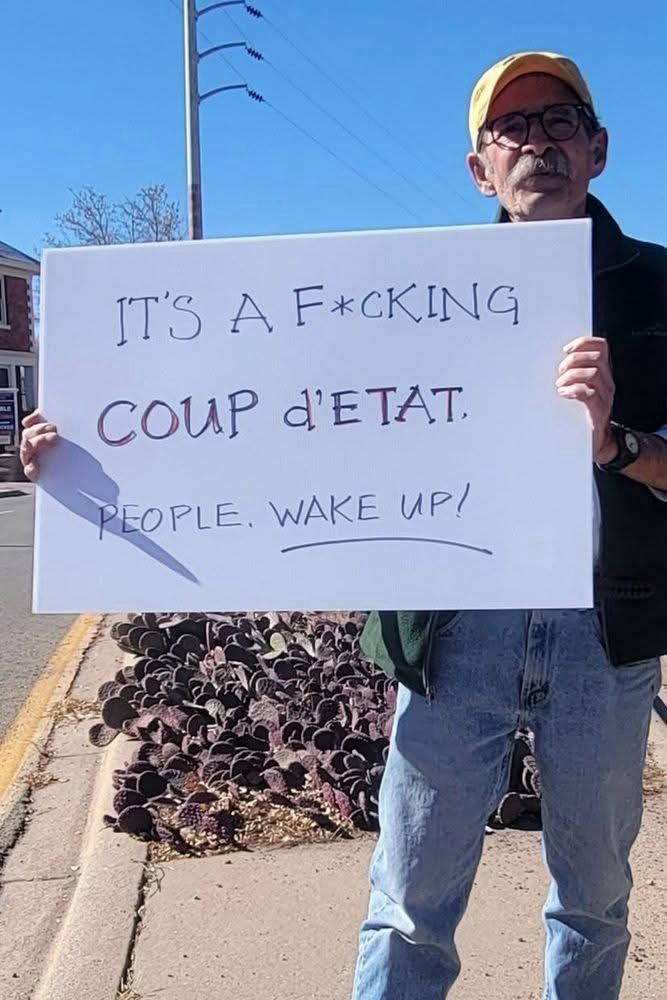 Man holding sign that reads: It’s a fucking coup d’etat. People wake up!