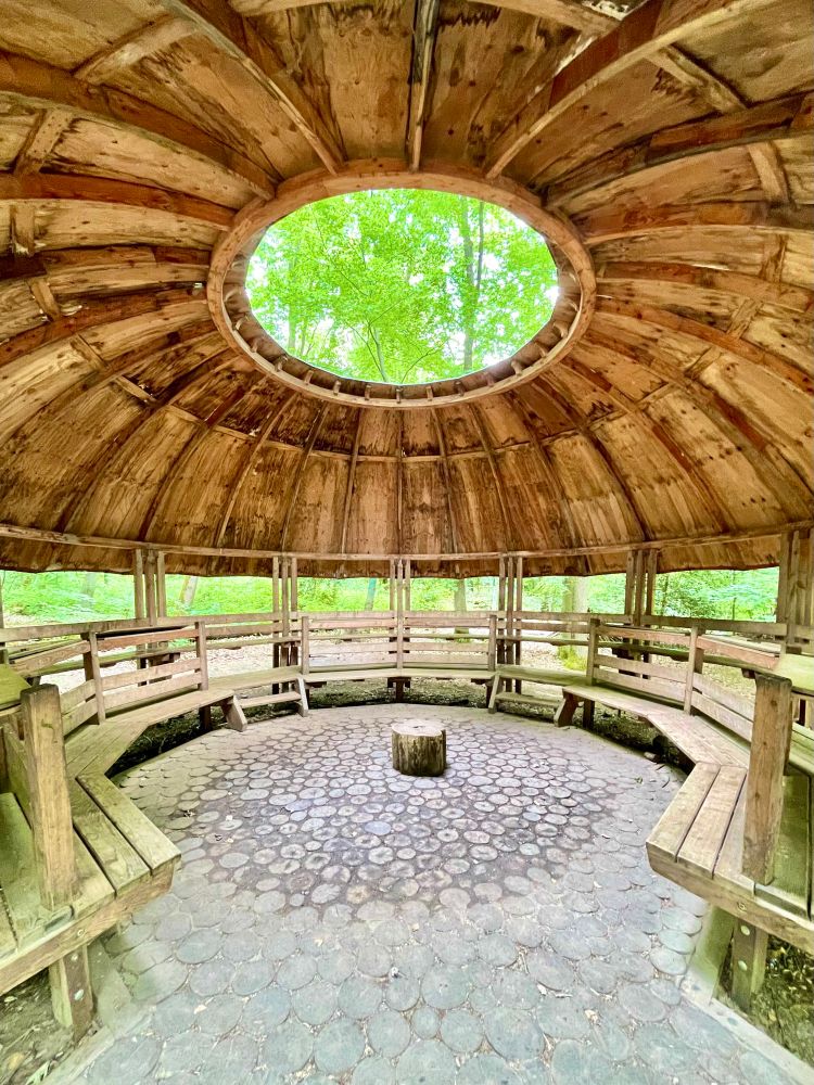 Das Foto zeigt eine runde, offene Holzhütte, die sich mitten im Wald im Wildpark Saarbrücken befindet. Die Konstruktion ist aus natürlichen, unbehandelten Holzplanken gebaut, was ihr ein rustikales, naturverbundenes Aussehen verleiht. Das Dach ist ebenfalls rund und wird von einem breiten, offenen Kreis in der Mitte unterbrochen – wie ein Auge zum Himmel. Durch diese Öffnung fällt Tageslicht ins Innere, und man kann die Baumwipfel und den blauen Himmel sehen.

Innen zieht sich eine kreisförmige Holzbank an der Wand entlang, sodass sich Besucher rundherum hinsetzen können. In der Mitte des Hüttenkreises steht ein kleiner Baumstumpf auf einem Boden aus kreisrund geschnittenen Holzscheiben. Zwischen den Holzplanken der Wand sind Spalten, durch die man das satte Grün des umgebenden Waldes erkennen kann.