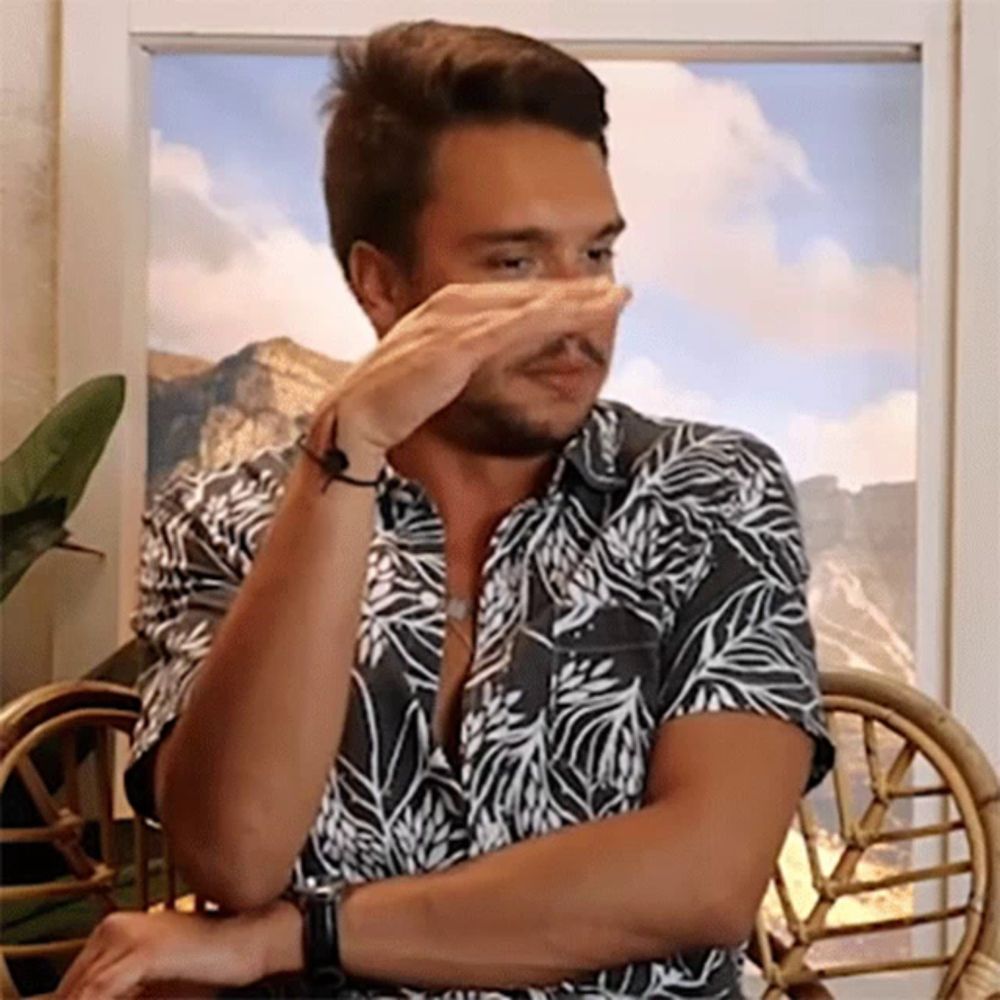 a man in a hawaiian shirt is covering his nose with his hand while sitting in a chair .