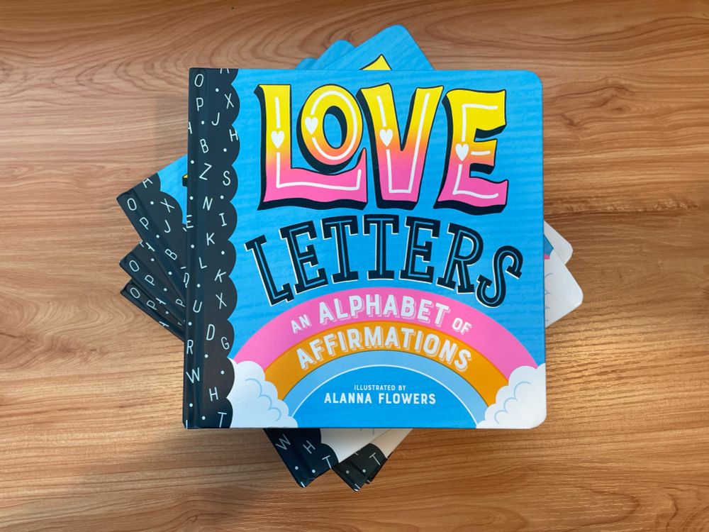 A stack of the book “Love Letters: An Alphabet of Affirmations” on a wood desk