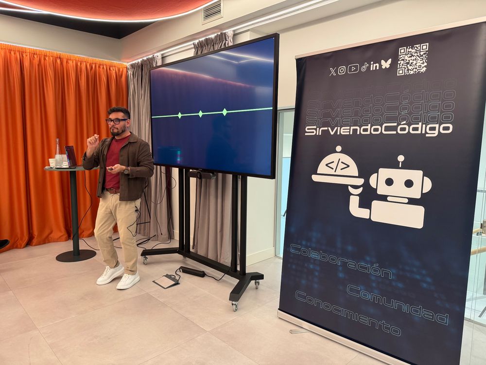 Una persona expone en una sala, delante de una pantalla. Está de pie.