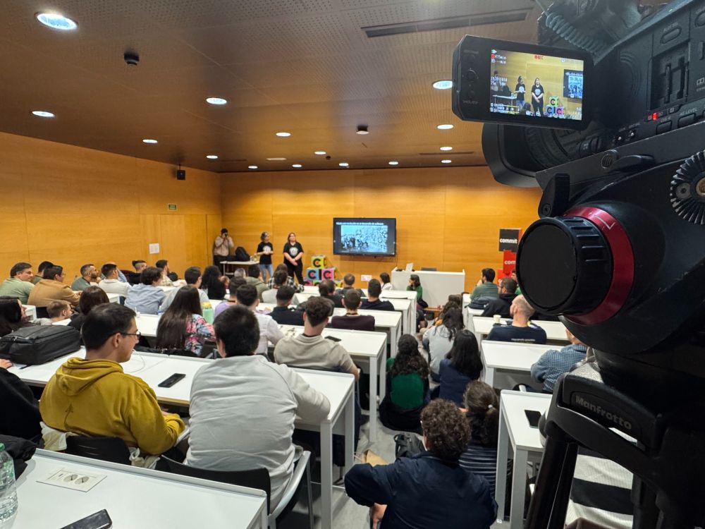 Se ve una sala de conferencias. Al fondo, dos personas exponen al lado de una pantalla de televisor. En primer plano, se ve una parte de una cámara de vídeo profesional con la pantalla de previsualización desplegada. Entre la cámara y las personas que exponen, hay mesas con personas sentadas. También aparecen personas sentadas en el suelo. 