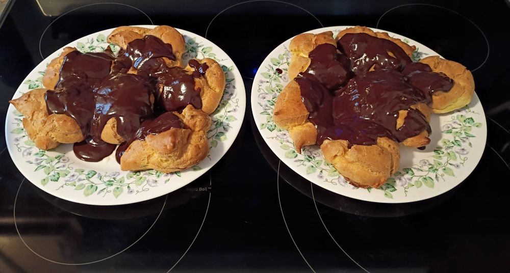 Two large, decorative white plates hold 12 profiteroles, which have been filled with cream and smothered in melted chocolate. 