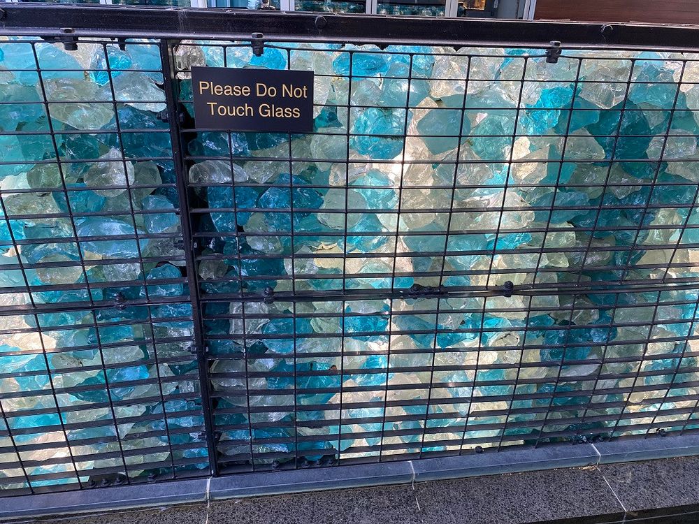 Close-up picture of glass "sculpture" consisting of chunks of glass held behind wire. A sign on it says "Please Do Not Touch Glass".