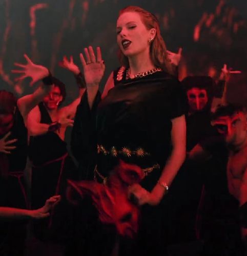 From the Karma music video: Taylor Swift dressed in black with gold chain accents holding a horned mask in one hand and waving with the other, standing in front of a group of people dressed in black hoods and creepy masks under red lighting.