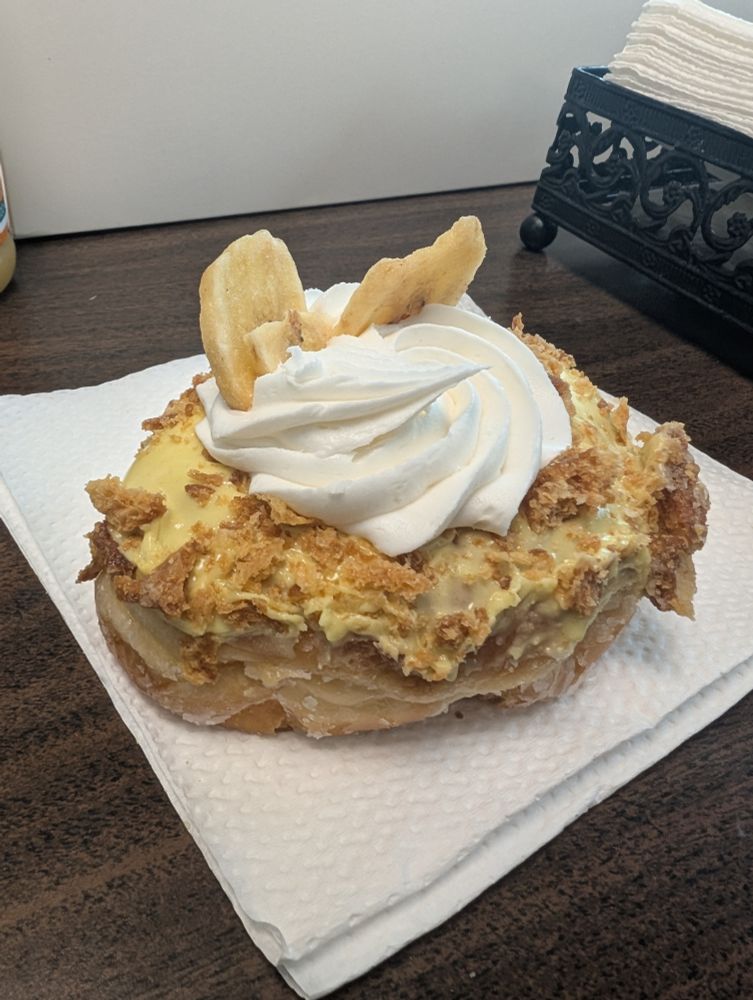 A table with a napkin and a donut with yellow frosting and crumbles on top and a dollop of white cream in the middle with two banana chips sticking out of the top.