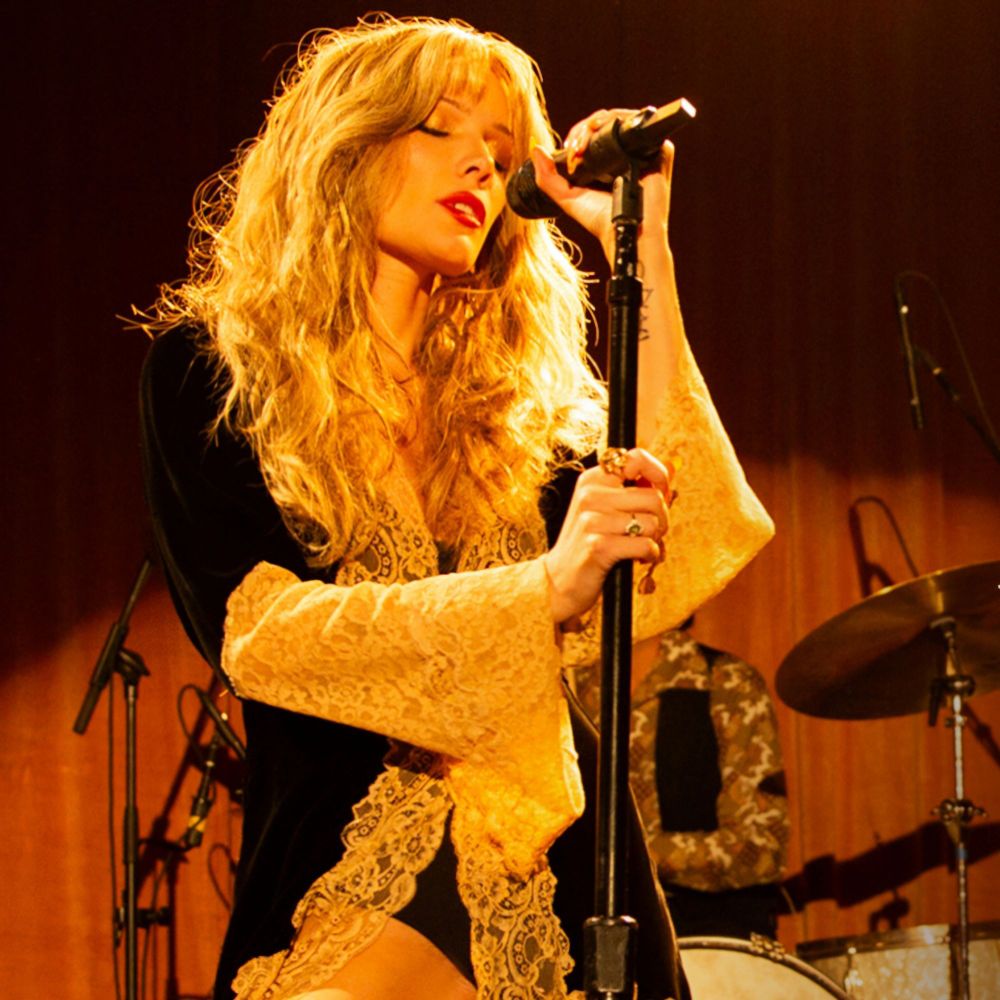 Halsey with long blonde hair wearing a black robe with lacey trim and bell sleeves. She's holding onto a mic stand singing with a drum set and red curtain behind her.