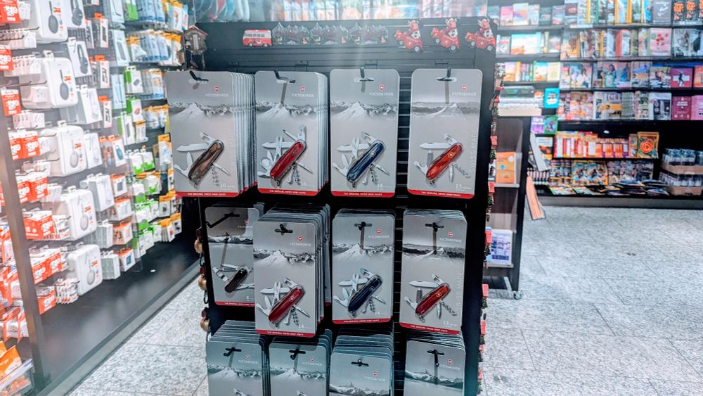 Photo of a large retail display in a shop inside Zurich airport.  Amid the variety of snacks and technology accessories like earbuds and charging cables we also see a large rack of Swiss Army Knives.  These are models that even include blades.