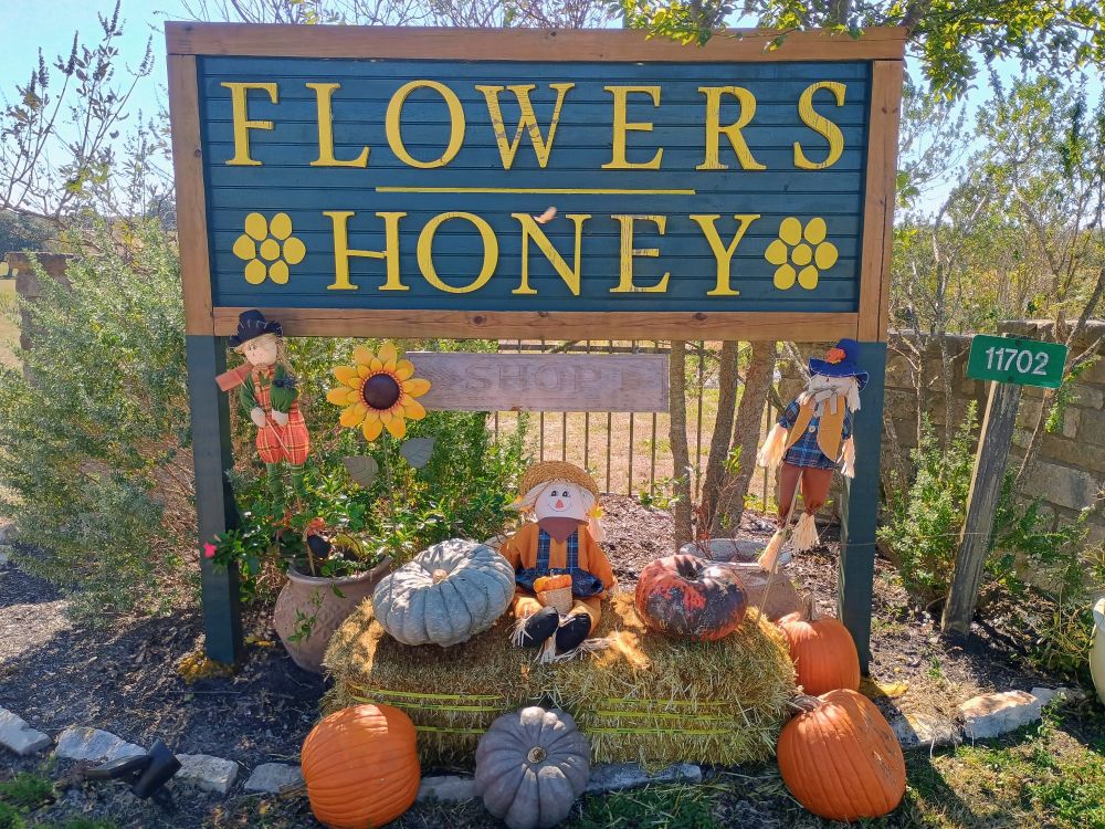 A blue and yellow sign for a store called "Flowers and Honey". Underneath the outdoor sign, pumpkins and scarecrow dolls decorate the ground. Trees and lilacs are in the background.