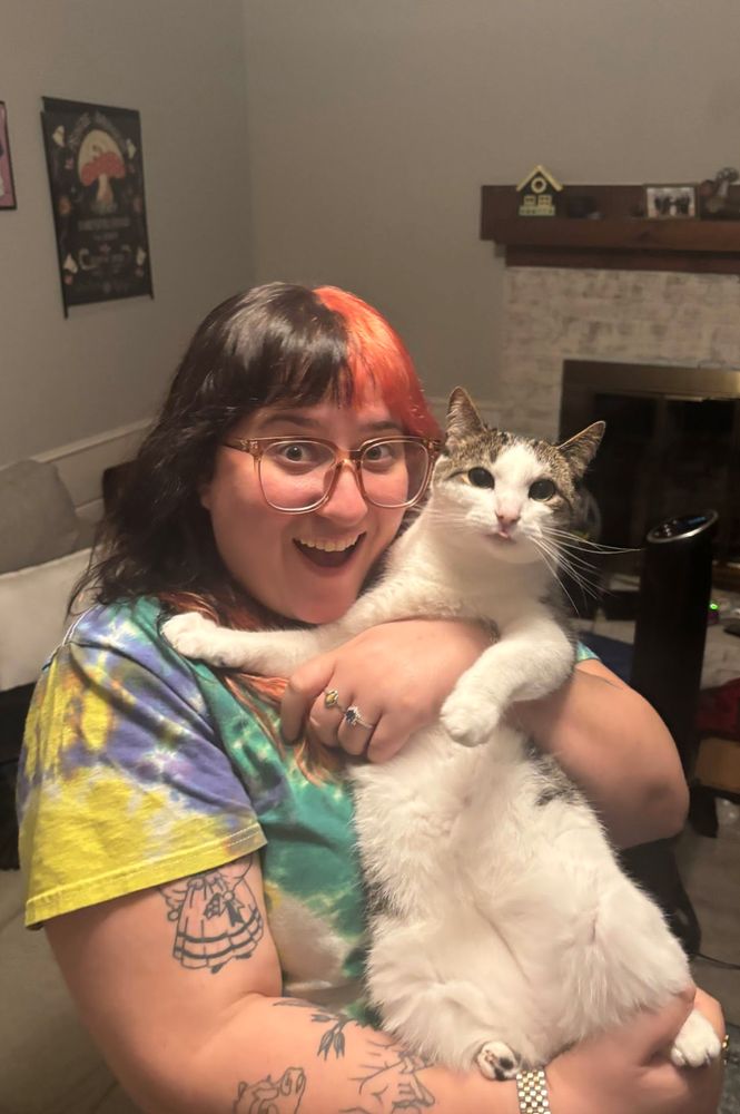 a woman with pink and brown hair holding a white and grey cat who is sticking his tongue out