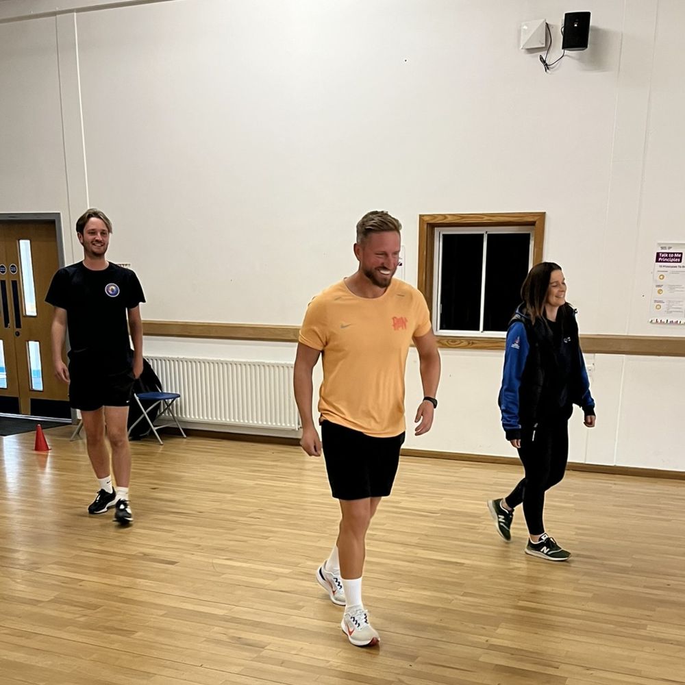Three people walking across the wooden floor of a sports hall. The person on the left is wearing a black t-shirt, black shorts and black running shoes. The person in the middle is wearing an orange t-shirt, black shorts and white running shoes. The person on the right is wearing a black and blue hoodie, black leggings and black running shoes.