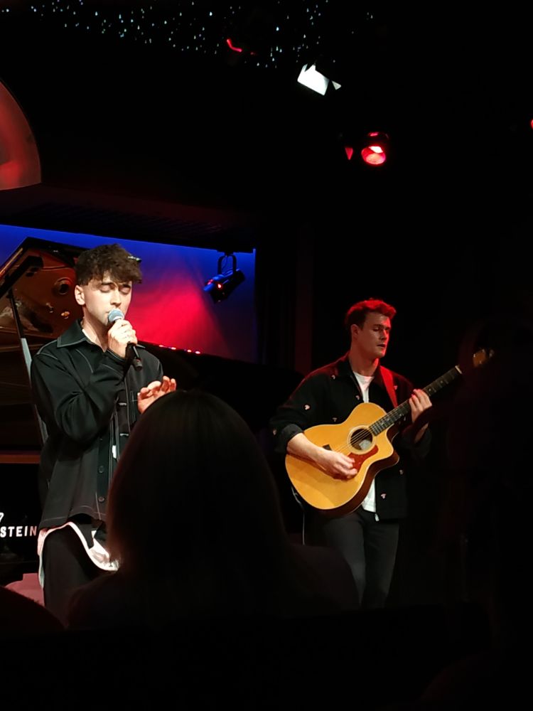 Benedict is standing on the left, singing with his eyes closed, while Lloyd is playing the guitar on the right side corner.