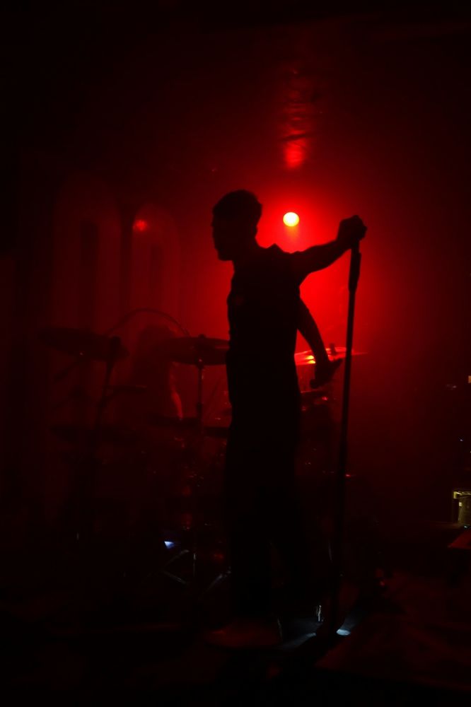 Dan Smith silhouette emerge from a red background, holding the foot of his microphone. He seems to look back just before facing us again to continue the concert.
