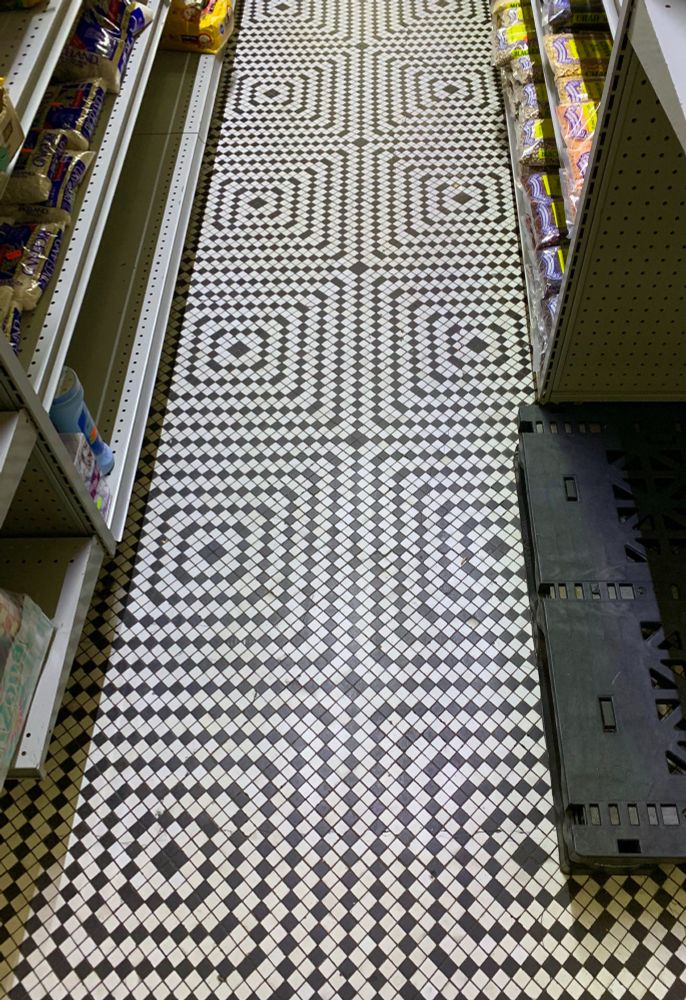 A narrow aisle of the neighborhood corner store has an aged black & white tile floor.
