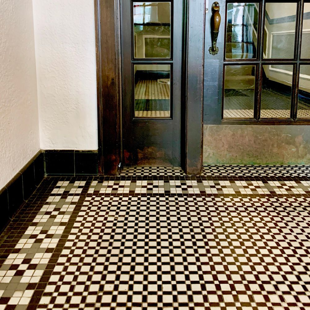 A circa 1920 apartment building entrance hallway with dark wood trim, glass door and side light, curved door handle and black, white and grey mosaic tile floor. 