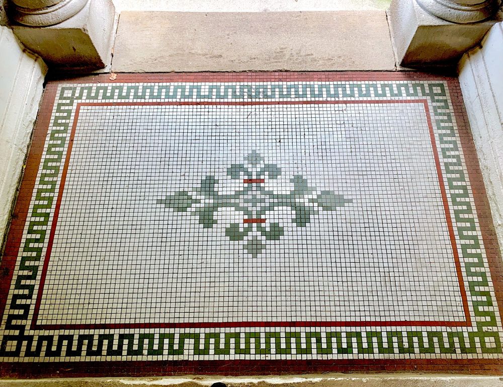 A white vintage tile floor with two small red bars over a green center flourish, a green meander border outlined in red and the base of two concrete columns at the entrance of an apartment building.