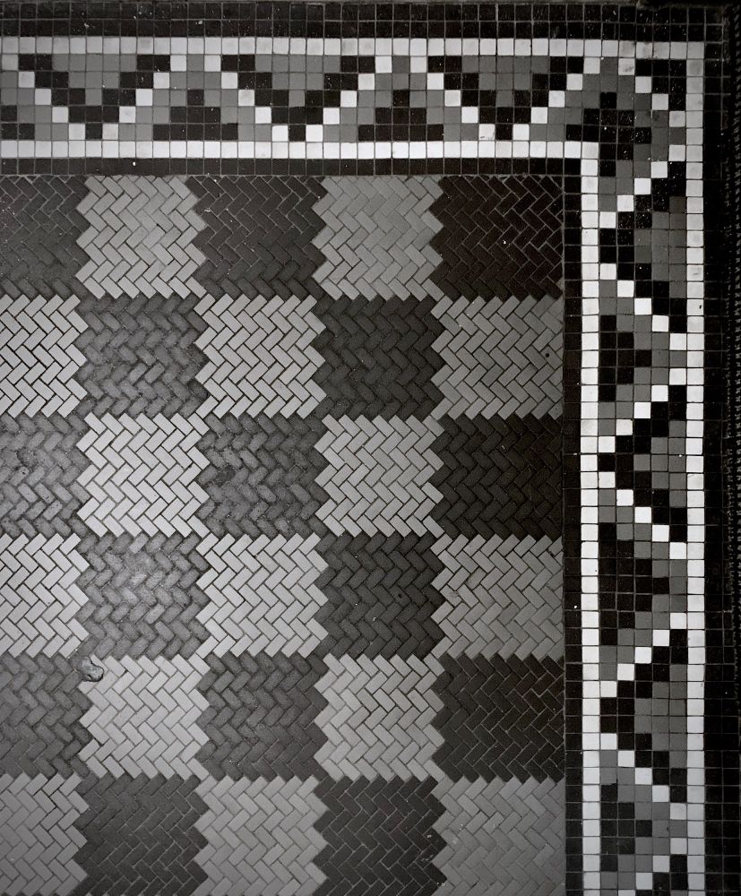 The corner of a black and grey vintage tile floor made from small rectangular tiles in the herringbone pattern and a border with small square tiles making a zig zag pattern in white, grey and black.