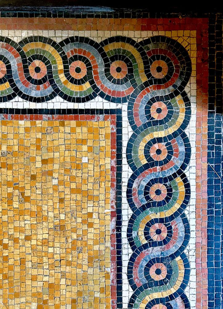 A vintage mosaic tile floor with a center field of rust colored tiles and a colorful boarder with a double loop meander in black, green, rust, blue, red, orange and white.