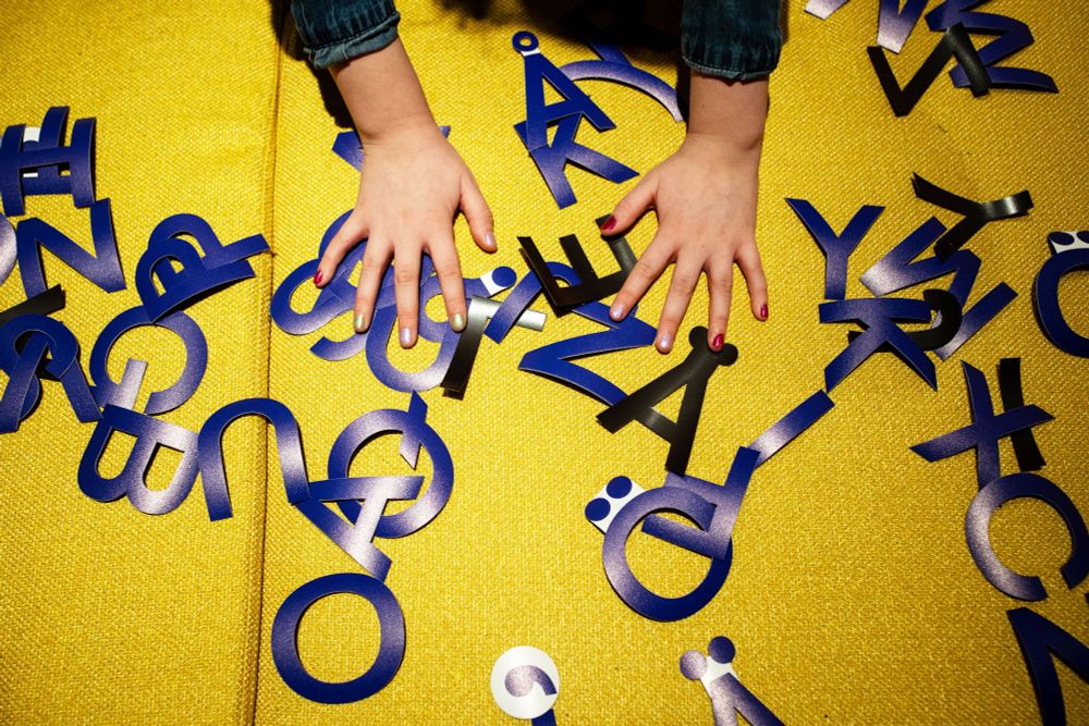 Sinisiä magneettikirjaimia keltaisella penkillä. Kirjainten päällä kädet, jotka siirtelevät kirjaimia.