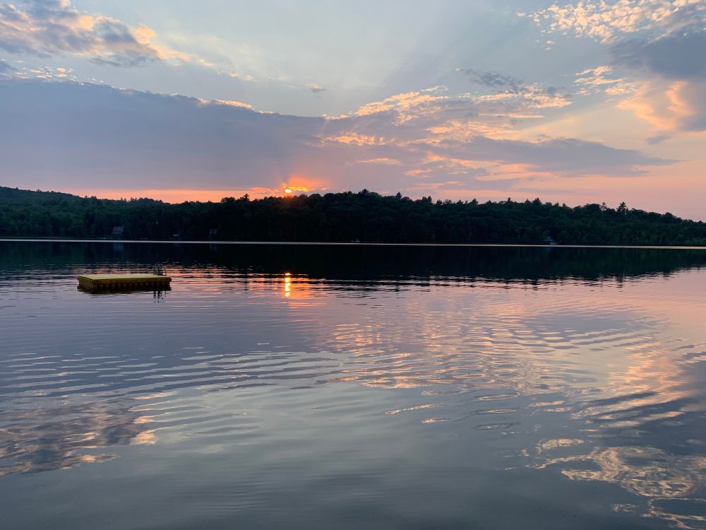 photo of a sunset across and reflected on a still pond surrounded by trees with clouds in the sky and a swimming float in the foreground