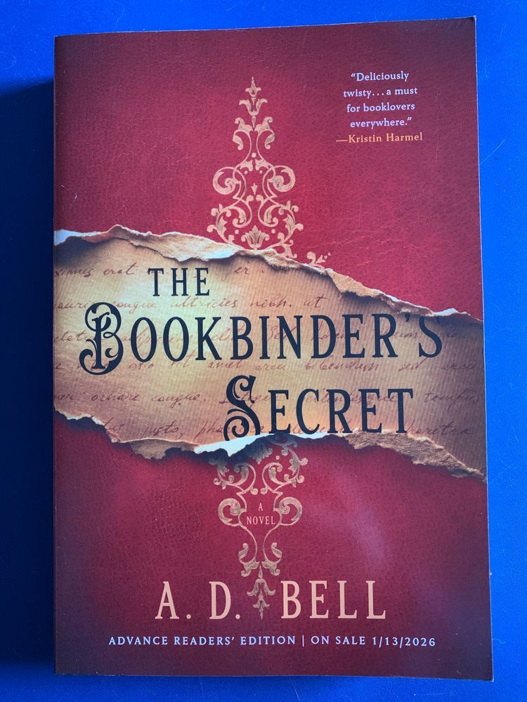 book cover for the bookbinder’s secret by a. d. bell with black lettering over a textured red and gold background evoking an antique book cover with the title in a troupe i’oeil tear through the middle of the cover 