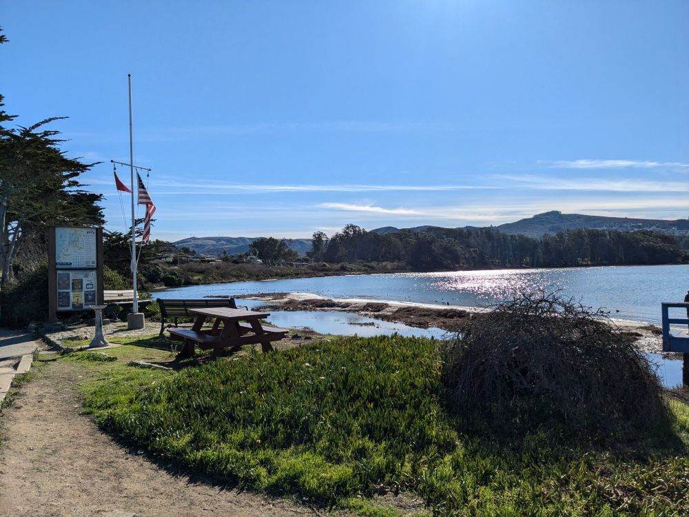 A bay of water on a sunny day. There's are trees and hills in the background. In the foreground is an empty picnic table.
