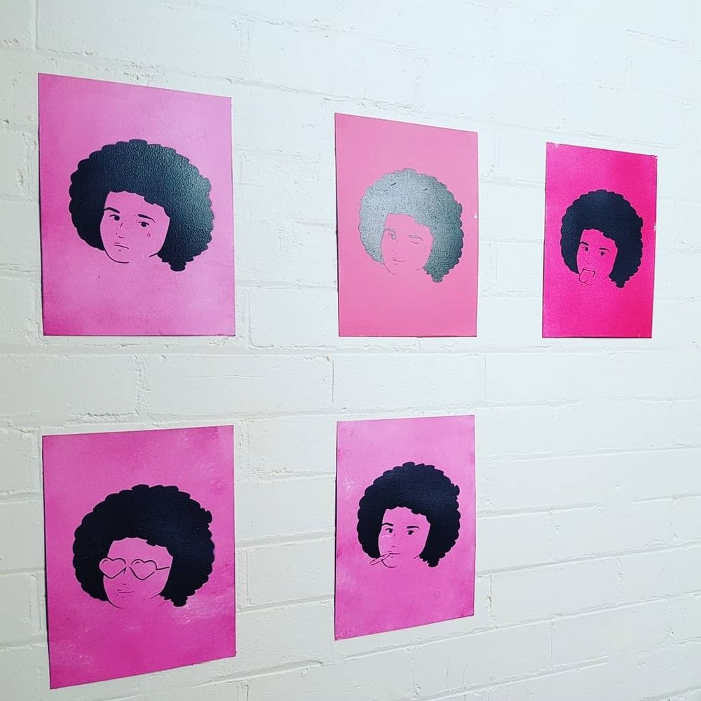 Five pink canvas panels hung on a white brick wall, each depicts a woman with an afro and slight variations of facial expression. Left to right top to bottom are: crying, winking, tongue out, with loveheart shaped glasses, and smoking a joint