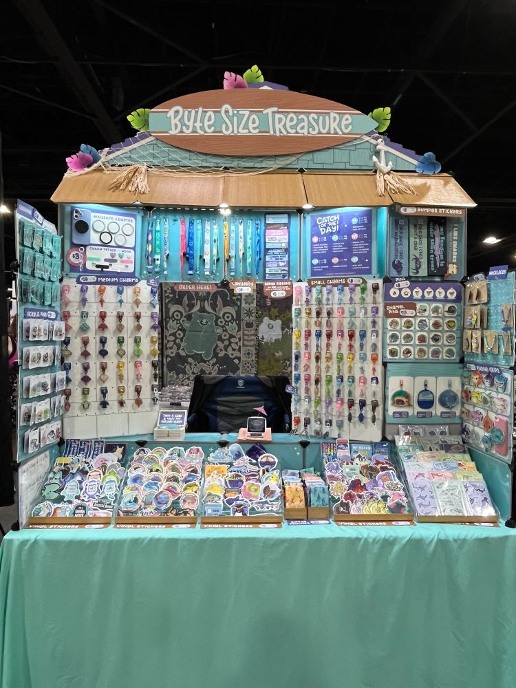 Convention artist booth that looks like a boardwalk shop, full of colorful shark & fish merchandise. From stickers, charms, to bags, and jewelry! 