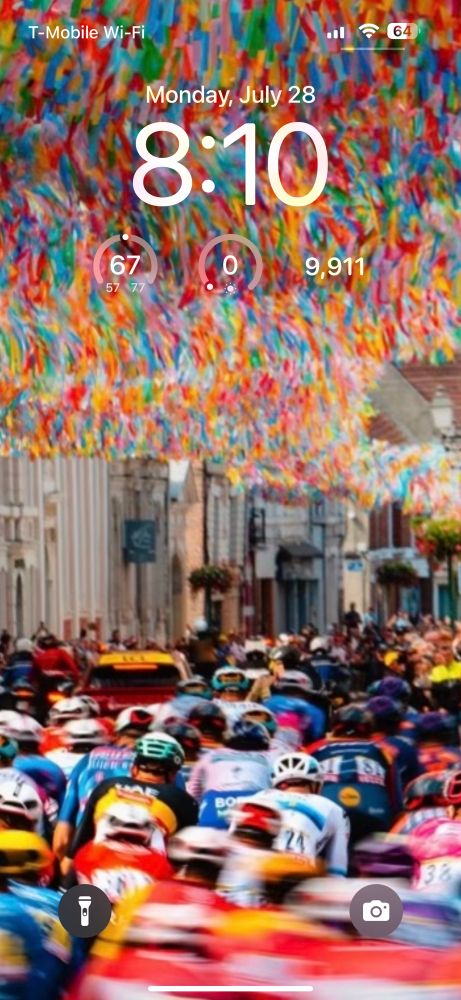 Lock Screen showing the riders of the Tour de France going through a town center canopied with rainbow streamers 