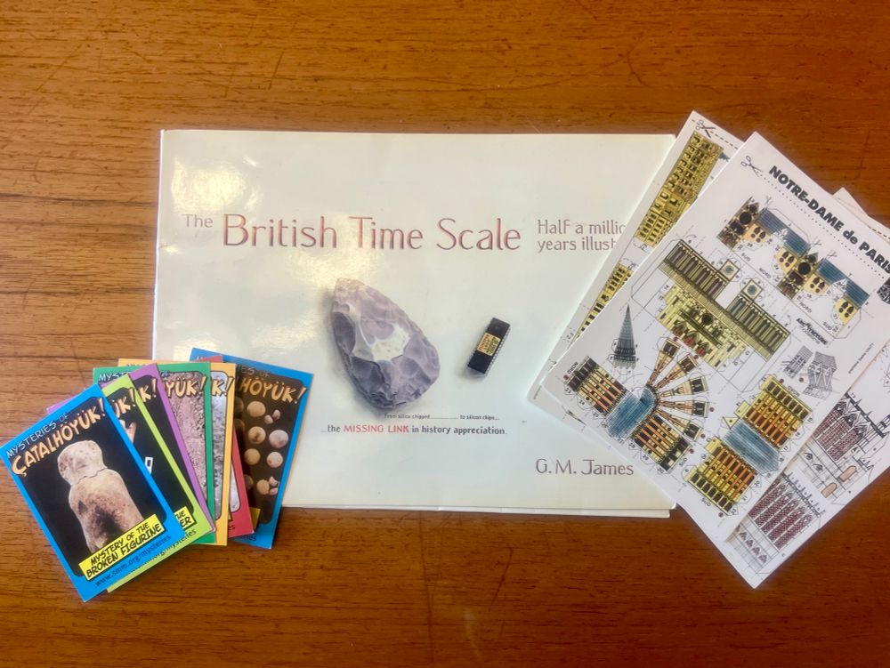 A collection of paper products for archaeological education and outreach. On the left are several cards fanned out. The cards are in a comic book trading card style, each with a picture of an artifact from Çatalhöyük and a brightly colored border. On the right are 3 postcards from Paris, France that can be cut up to create paper models of famous architecture. The top card is for Notre-Dame. A thin booklet, titled “The British Time Scale: Half a million years illustrated”, is in the center. 