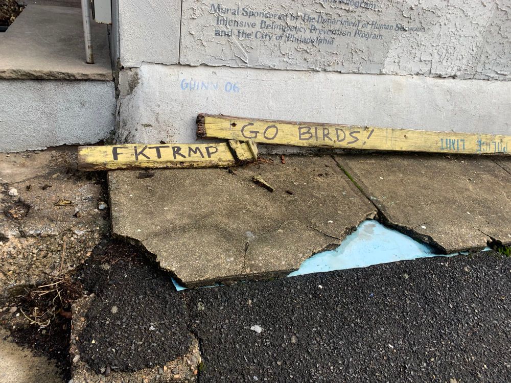 Broken wooden police barriers on the ground beneath a mural, one piece says FKTRMP the other piece says GO BIRDS!