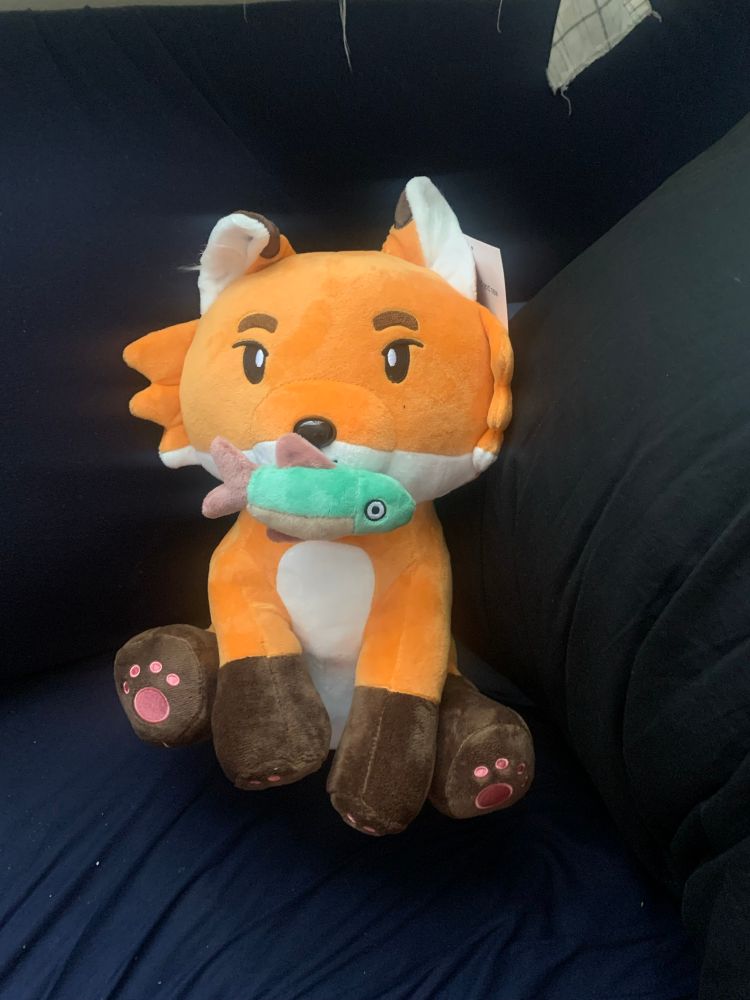 A fox plush from the podcast Worlds Beyond Number, there is a magnet fish at his mouth and one ear is bent forward to make him look curious and rakish 