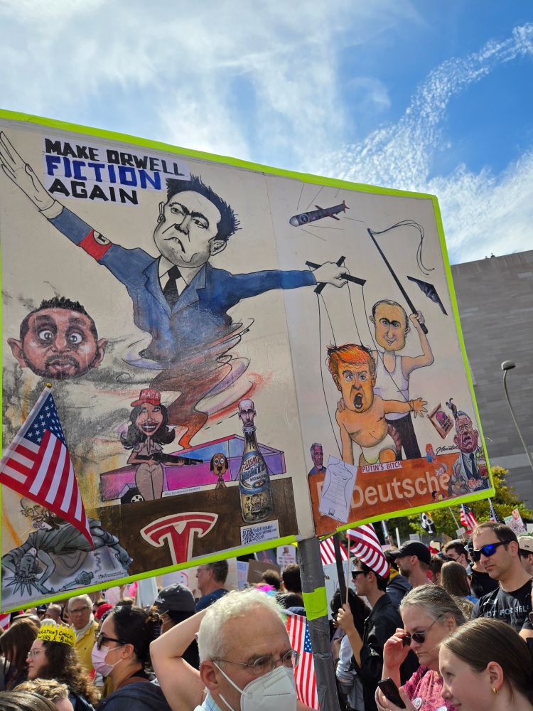 A protest sign that reads "Make Orwell Fiction Again" and shows cartoon drawings of Elon Musk with an SS armband and controlling Trump as a baby marionette. Also features Kash Patel, Stephen Miller, and Putin with a whip. 