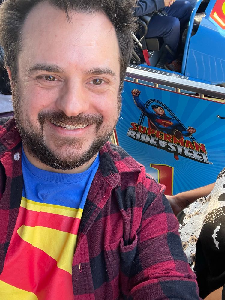 A high-angle selfie of a smiling, bearded 37-year-old man wearing a Superman (2025) logo shirt under a red flannel shirt. In the background is the row 1 gate for Superman: Ride of Steel at Six Flags America. The ride car is just barely visible right behind the gate.