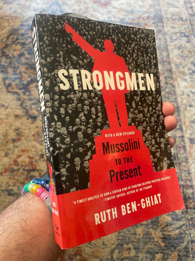A man wearing a couple of plastic bead bracelets, holding a paperback copy of Ruth Ben-Ghiat’s book “Strongmen: Mussolini to the Present”.