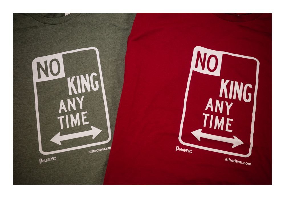 Two t-shirts lay side by side, one olive green and one deep red, featuring a bold white graphic that mimics a New York City parking sign. The design reads “NO KING ANY TIME” with double-headed arrows pointing left and right at the bottom. Below are the logos for BetaNYC and alfredtwu.com.