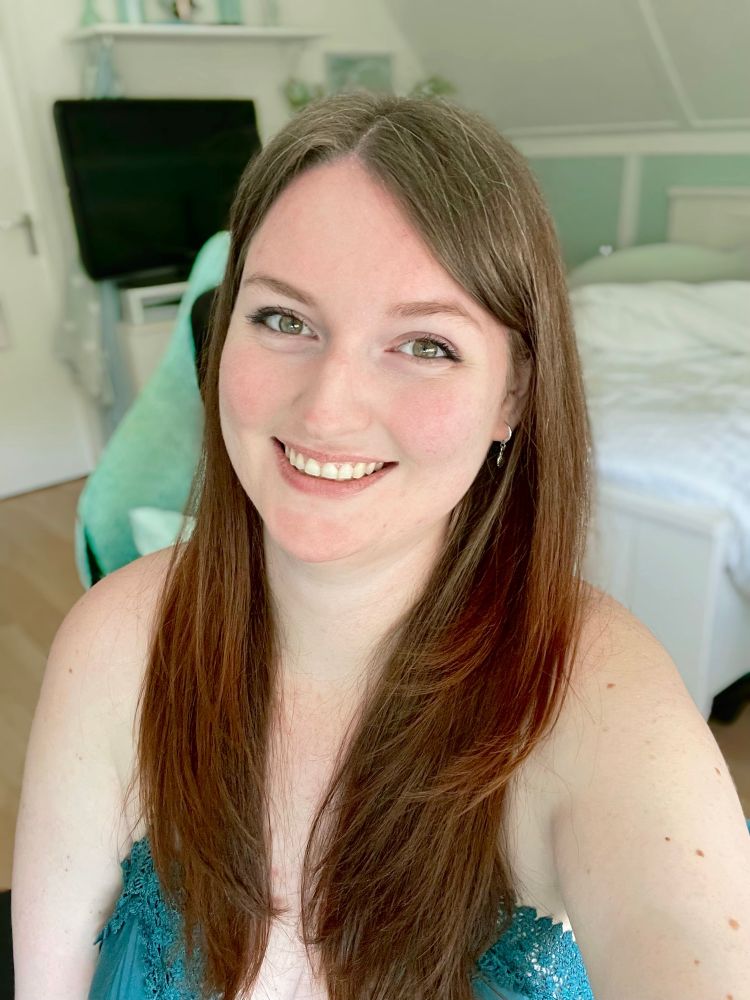 It’s me! I am a young woman with reddish brown hair and hazel/green eyes. In this selfie, I’m smiling widely for the camera, and I’m wearing a teal summer dress. You can see my bedroom in the background, though it is out of focus.