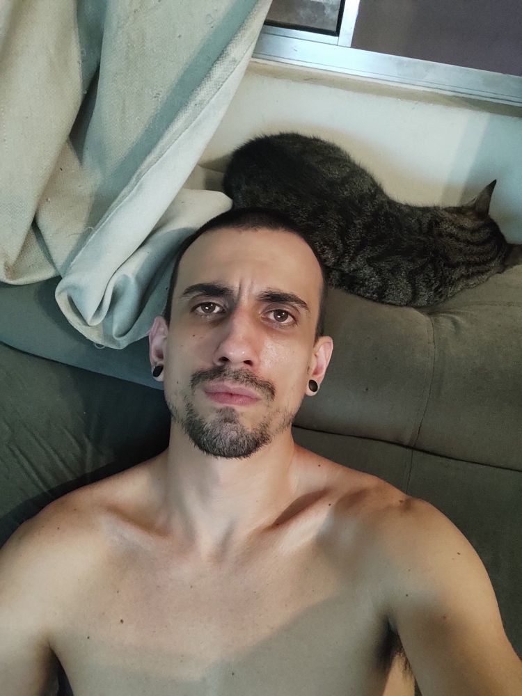 Eu, uma pessoa latino-americana com cabelo raspado curto, barba por fazer e alargadores nas orelhas, sentado no sofá com meu gato dormindo ao fundo