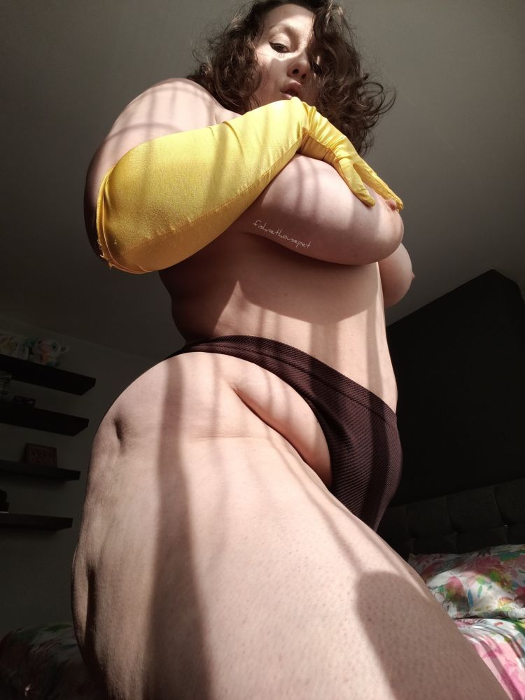 Fishnethousepet posed from the side and above, wearing a brown thong and yellow opera glove. They look down at you as they caress their big tits