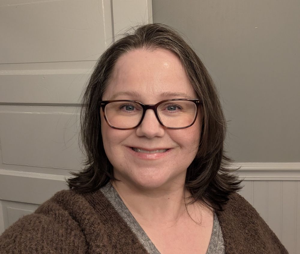 A smiling woman wearing a brown cardigan over a gray T-shirt with brown-framed glasses on.