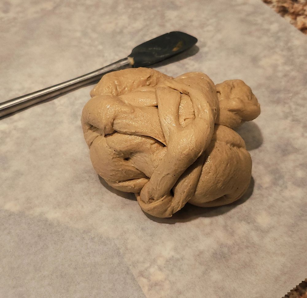 A knotted ball of seitan on parchment paper. Next to the seitan is a mixing spatula for scale. The spatula is metal and silicone. 